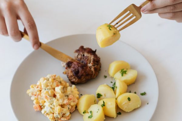 From above of crop anonymous person with cutleries eating delicious fried cutlet and hot boiled potatoes with parsley garnished with nutritious Russian salad